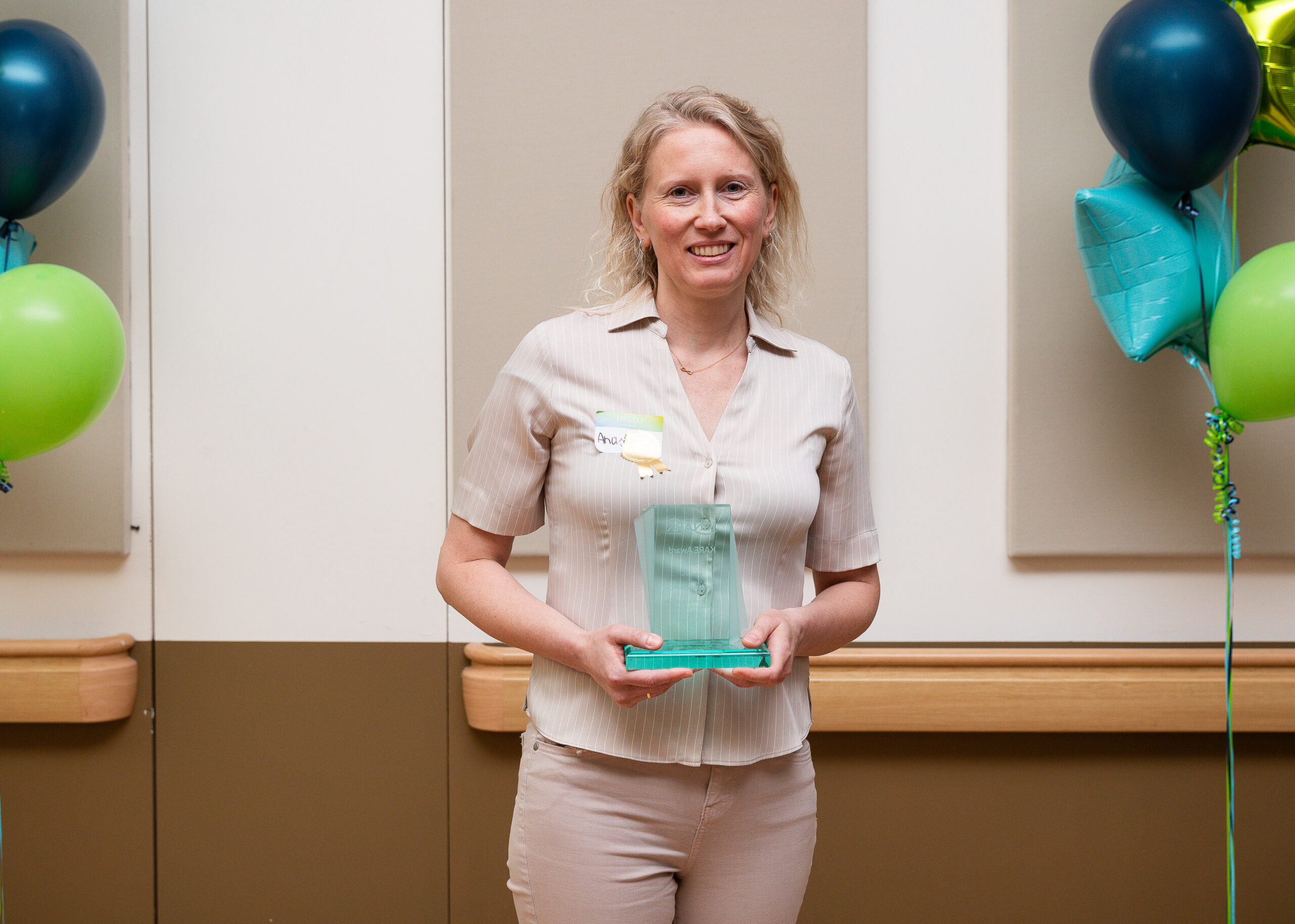 A photo of a woman with blonde hair and a cream coloured top smiling and holding a glass KARE Award in front of balloons. 