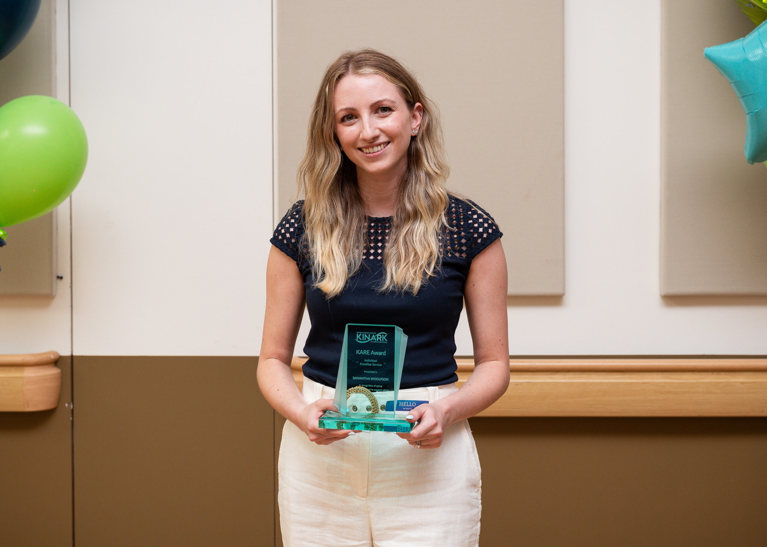 A photo of a woman with blonde hair and a navy shirt, smiling and holding a glass KARE Award in front of balloons. 