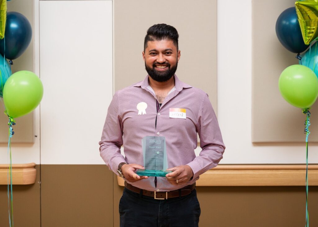 A photo of a man in a button up shirt smiling and holding a glass KARE Award in front of balloons. 